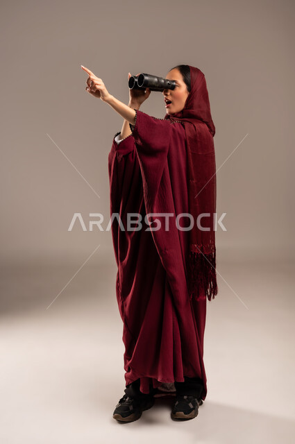 رؤية الاشياء البعيدة عبر التلسكوب، التخطيط المستقبلي للأهداف و الطموحات، حب الاستكشاف والمغامرة، بورتريه لامرأة عربية خليجية سعودية محجبة ترتدي العباءة الحمراء تنظر من خلال منظار ثنائي العينين, الجسم بالطول الكامل، خلفيه بيج