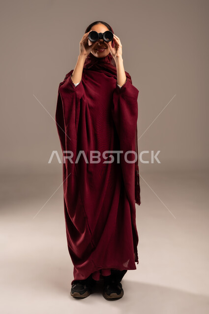 Seeing distant objects through a telescope, future planning for goals and ambitions, love of exploration and adventure, portrait of a veiled Saudi Arabian Gulf woman wearing a red abaya looking through binoculars, full body, beige background