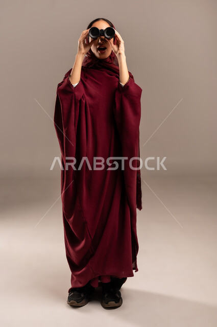 رؤية الاشياء البعيدة عبر التلسكوب، التخطيط المستقبلي للأهداف و الطموحات، حب الاستكشاف والمغامرة، بورتريه لامرأة عربية خليجية سعودية محجبة ترتدي العباءة الحمراء تنظر من خلال منظار ثنائي العينين, الجسم بالطول الكامل، خلفيه بيج