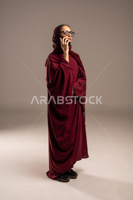 مشاركة الأخبار والأحاديث, التواصل مع العائلة والأصدقاء, استخدام الأجهزة التقنية الحديثة في الحياة اليومية, بورتريه لامرأة عربية خليجية سعوديه محجبة ترتدي العباءة الحمراء تقوم بإجراء اتصال هاتفي عبر الهاتف المحمول, الجسم بالطول الكامل، خلفية بيج