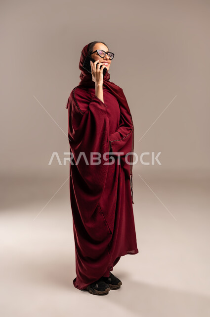 مشاركة الأخبار والأحاديث, التواصل مع العائلة والأصدقاء, استخدام الأجهزة التقنية الحديثة في الحياة اليومية, بورتريه لامرأة عربية خليجية سعوديه محجبة ترتدي العباءة الحمراء تقوم بإجراء اتصال هاتفي عبر الهاتف المحمول, الجسم بالطول الكامل، خلفية بيج