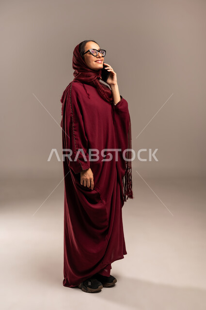 مشاركة الأخبار والأحاديث, التواصل مع العائلة والأصدقاء, استخدام الأجهزة التقنية الحديثة في الحياة اليومية, بورتريه لامرأة عربية خليجية سعوديه محجبة ترتدي العباءة الحمراء تقوم بإجراء اتصال هاتفي عبر الهاتف المحمول, الجسم بالطول الكامل، خلفية بيج
