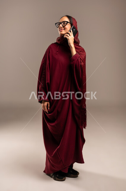Sharing news and conversations, communicating with family and friends, using modern technology in daily life, portrait of a veiled Saudi Arabian Gulf woman wearing a red abaya making a phone call via mobile phone, full body, beige background