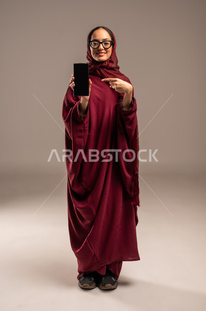 استخدام الأجهزة التقنية الحديثة, النظر إلى الكاميرا بإيماءات السعادة و السرور, بورتريه  لشابة عربية خليجية سعودية مبتسمة محجبة ترتدي العباءة الحمراء تشير باصبع السبابة للهاتف المحمول، عرض شاشة سوداء فارغة, الجسم بالطول الكامل، خلفيه بيج