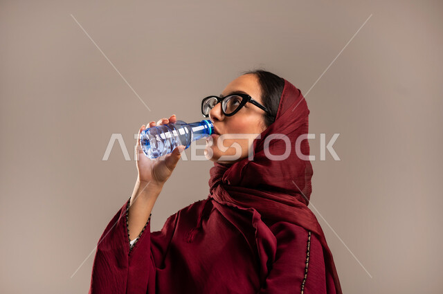 الارتواء بالماء للتخفيف من حراره الجو, الشعور بالعطش والظما, بورتريه مقرب لسيدة عربية خليجية سعوديه ترتدي العباءه الحمراء تقوم بشرب المياه من زجاجة ماء بلاستيكية صغيرة، المداومة على شرب المياه، المحافظة على سلامة الجهاز المناعي للجسم، خلفية بيج