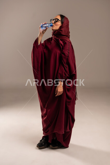 الارتواء بالماء للتخفيف من حراره الجو, الشعور بالعطش والظما, بورتريه لسيدة عربية خليجية سعوديه ترتدي العباءه الحمراء تقوم بشرب الماء من عبوة بلاستيكية صغيرة، المداومة على شرب المياه، المحافظة على سلامة الجهاز المناعي للجسم، الجسم بالطول الكامل،  خلفية بيج