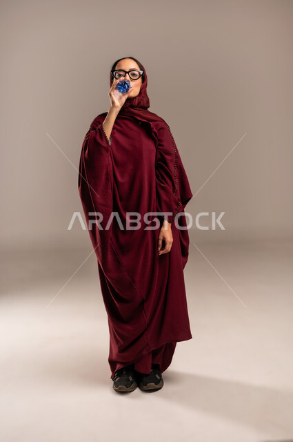 الارتواء بالماء للتخفيف من حراره الجو, الشعور بالعطش والظما, بورتريه لسيدة عربية خليجية سعوديه ترتدي العباءه الحمراء تقوم بشرب الماء من عبوة بلاستيكية صغيرة، المداومة على شرب المياه، المحافظة على سلامة الجهاز المناعي للجسم، الجسم بالطول الكامل،  خلفية بيج