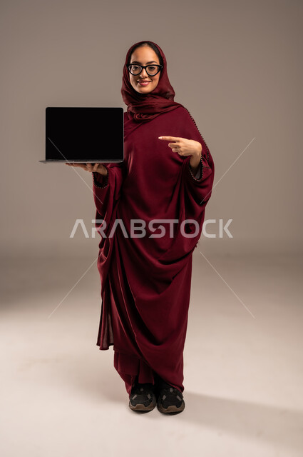 استخدام الاجهزة التقنية الحديثة، بورتريه مقرب لامرأة عربية خليجية سعودية محجبة ترتدي العباءه الحمراء تحمل بيدها جهاز الاب توب بشاشة سوداء وتشير بأصبع السبابة اليه، إيماءات السعادة والسرور، عرض تطبيقات وخدمات الهاتف، خلفية بيج