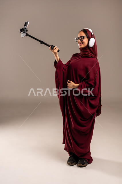 استخدام تقنية الهيدفون الحديثة, الاندماج و التفاعل مع الأنغام الموسيقية, الاستمتاع بسماع الأغاني المفضلة, بورتريه لامرأة عربية خليجية سعوديه محجبة ترتدي العباءة الحمراء تضع سماعات الرأس الصوتية وتقوم بالتقاط سيلفي, خلفية بيج