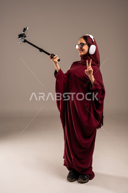 استخدام تقنية الهيدفون الحديثة, الاندماج و التفاعل مع الأنغام الموسيقية, الاستمتاع بسماع الأغاني المفضلة, بورتريه لامرأة عربية خليجية سعوديه محجبة ترتدي العباءة الحمراء تضع سماعات الرأس الصوتية وتقوم بالتقاط سيلفي, خلفية بيج