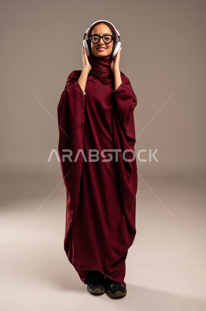 Merging and interacting with musical melodies, using modern headphone technology, enjoying listening to favorite songs, portrait of a veiled Saudi Arabian Gulf woman wearing a red abaya, putting audio headphones around her head and listening to music, full body, beige background