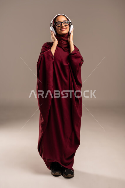 الاندماج و التفاعل مع الأنغام الموسيقية, استخدام تقنية الهيدفون الحديثة, الاستمتاع بسماع الأغاني المفضلة, بورتريه لامرأة عربية خليجية سعوديه محجبة ترتدي العباءة الحمراء تضع سماعات الرأس الصوتية حول رأسها وتقوم بالاستماع الى الموسيقى, الجسم بالطول الكامل، خلفية بيج