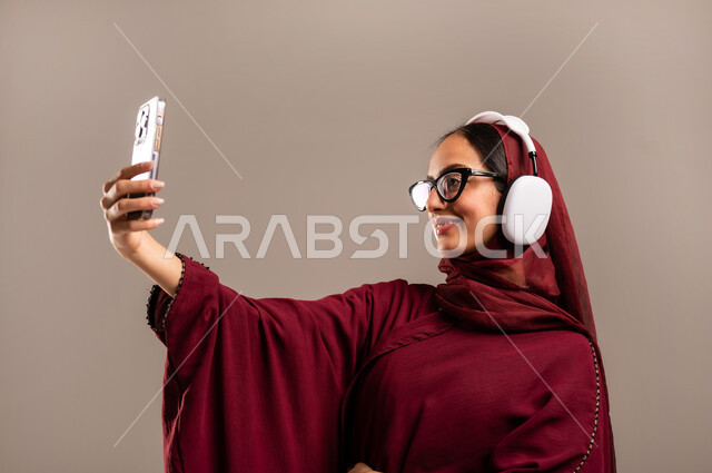 استخدام تقنية الهيدفون الحديثة, الاندماج و التفاعل مع الأنغام الموسيقية, الاستمتاع بسماع الأغاني المفضلة, بورتريه مقرب لامرأة عربية خليجية سعوديه محجبة ترتدي العباءة الحمراء تضع سماعات الرأس الصوتية وتقوم بالتقاط صوره سيلفي بهاتفهها المحمول, خلفية بيج