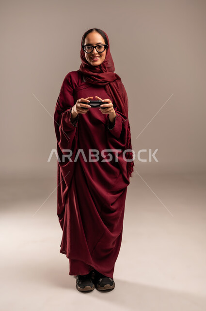 المتعة والتسلية بالفيديو جيمز، استخدام التقنيات الالكترونية الحديثة, بورتريه لإمرأة عربية خليجية سعوديه محجبة ترتدي العباءة الحمراء تحمل بيدها عصا التحكم بايماءات التفاعل والاندماج,  صورة الجسم بالكامل، خلفية بيج