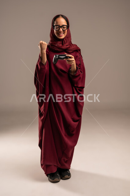 رفع قبضة اليد بتعابير الفوز والنصر، المتعة والتسلية بالفيديو جيمز، استخدام التقنيات الالكترونية الحديثة, بورتريه لإمرأة عربية خليجية سعوديه محجبة ترتدي العباءة الحمراء تحمل بيدها عصا التحكم بايماءات التفاعل والاندماج, صورة الجسم بالكامل، خلفية بيج