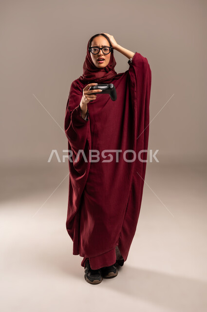 Expressions of loss and failure, fun and entertainment with video games, use of modern electronic technologies, portrait of a veiled Saudi Arabian Gulf woman wearing a red abaya holding a joystick with interaction and integration gestures, full body image, beige background