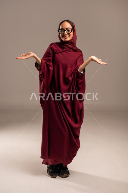 رفع اليدين للأعلى بإيماءات الامبالاه، بورتريه لامرأة عربيه خليجيه سعوديه محجبه ترتدي العباءة الحمراء ترفع يديها للاعلى بايماءات عدم تحمل المسؤوليه والاستهتار، عدم الاهتمام بأمر ما، الجسم بالطول الكامل، خلفية بيج