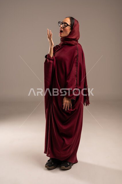 Raising hands up with yawning gestures, portrait of a veiled Saudi Arabian Gulf woman wearing a colorful abaya, putting her hand on her mouth with an expression of lethargy, sleepiness, and the need to rest and sleep, beige background