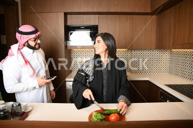 A Saudi Arabian Gulf woman preparing and preparing food, a Saudi Arabian Gulf man holding a mobile phone in his hand and talking to his wife while cutting vegetables, preparing delicious salads, preparing a healthy meal