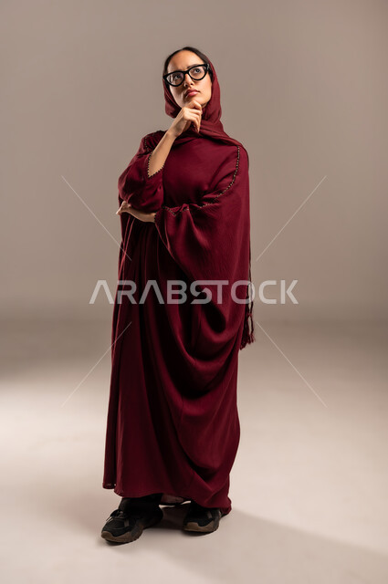 شرود الذهن في موضوع معين، النظر إلى شيء ما بإيماءات التأمل والتمعن, تعابير الاندماج و الحيرة , بورتريه لإمرأة عربية سعودية خليجية محجبة ترتدي العباءه الحمراء تضع يدها على رأسها بايماءات التفكير وايجاد الحلول لمشكلة ما، صورة الجسم بالطول الكامل، خلفية بيج