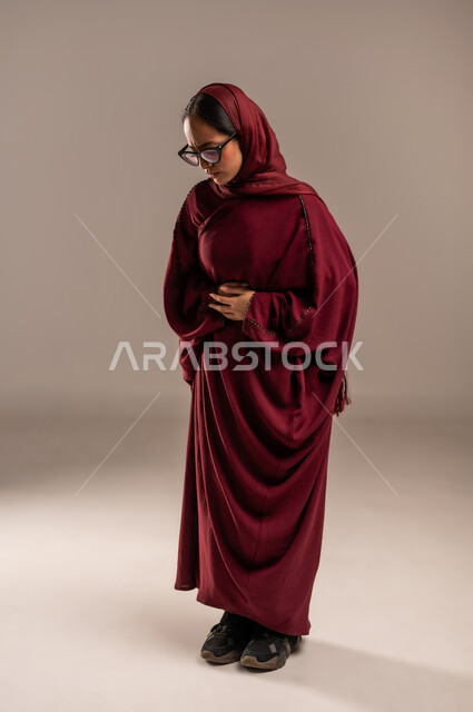 الشعور بحرقة في المعدة وعسر الهضم، الحاجه للمساعدة وطلب العلاج، الإحساس بالمغص والوجع، بورتريه من الجانب  لامرأه عربيه خليجيه سعوديه ترتدي عباءة حمراء تضع يدها على بطنها تنحني من شدة الألم، إيماءات التقيؤ والغثيان، صورة لكامل الجسم، خلفية بيج