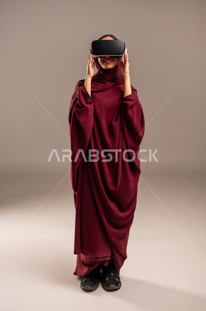 Expressions of surprise and delight, simulating a fantasy world, wearing virtual reality glasses, using a modern and advanced technical device for entertainment, close-up portrait of a Saudi Arabian Gulf woman wearing a hijab and a red abaya, wearing VR glasses, full-length body, beige background