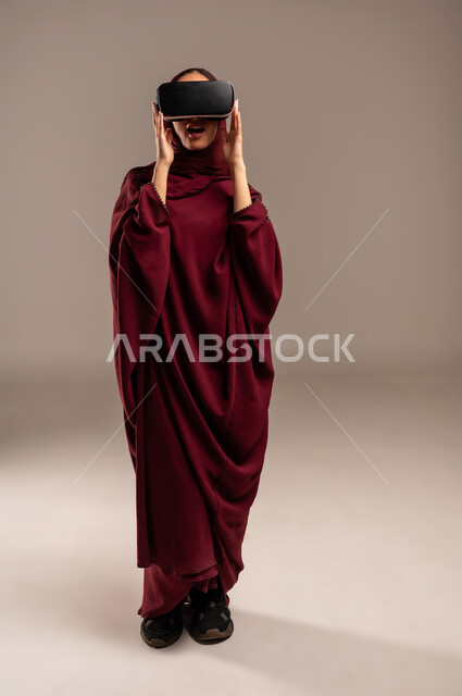 Expressions of surprise and delight, simulating a fantasy world, wearing virtual reality glasses, using a modern and advanced technical device for entertainment, close-up portrait of a Saudi Arabian Gulf woman wearing a hijab and a red abaya, wearing VR glasses, full-length body, beige background