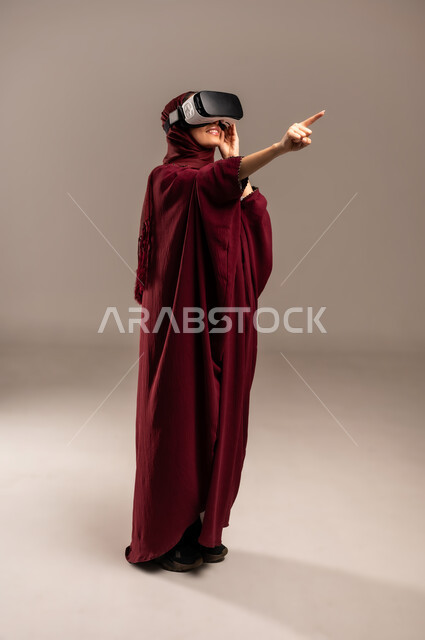 الاشارة الى شيء ما بتعابير الدهشة، محاكاة العالم الخيالي, ارتداء نظارة الواقع الافتراضي, استخدام جهاز تقني حديث ومتطور للتسلية والترفيه, بورتريه لسيده عربية خليجية سعودية ترتدي الحجاب والعباءة الحمراء تضع نظارات الفي ار, الجسم بالطول الكامل، خلفية بيج