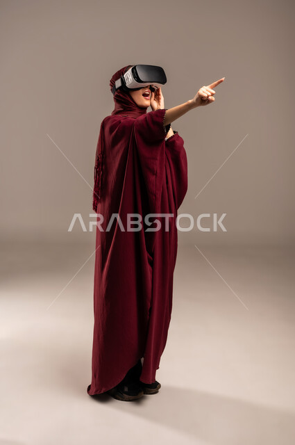 Pointing at something with an expression of surprise, simulating an imaginary world, wearing virtual reality glasses, using a modern and advanced technical device for entertainment, portrait of a Saudi Arabian Gulf woman wearing a hijab and a red abaya, wearing VR glasses, full body, beige background