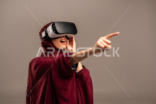 Pointing at something with an expression of surprise, simulating a fantasy world, wearing virtual reality glasses, using a modern and advanced technological device for entertainment, close-up portrait of a Saudi Arabian Gulf woman wearing a hijab and a red abaya, wearing VR glasses, beige background.
