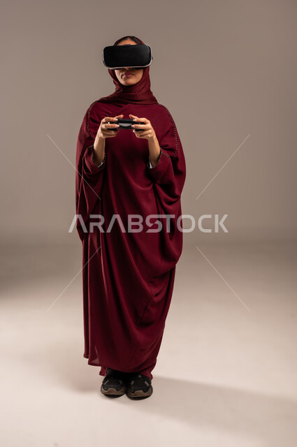 Gestures and expressions of enthusiasm and happiness, spending leisure time in video games, portrait of a Saudi Arabian Gulf woman wearing a hijab and a red abaya, wearing 3D VR glasses, holding a joystick in her hand and playing video games, full body, beige background