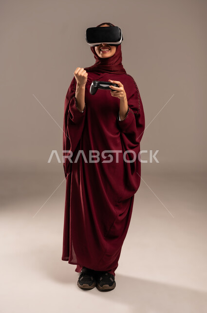 Gestures and expressions of enthusiasm and happiness, spending leisure time in video games, portrait of a Saudi Arabian Gulf woman wearing a hijab and a red abaya, wearing 3D VR glasses, holding a joystick in her hand and playing video games, full body, beige background
