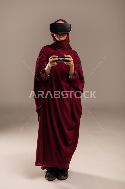 Gestures and expressions of enthusiasm and happiness, spending leisure time in video games, portrait of a Saudi Arabian Gulf woman wearing a hijab and a red abaya, wearing 3D VR glasses, holding a joystick in her hand and playing video games, full body, beige background