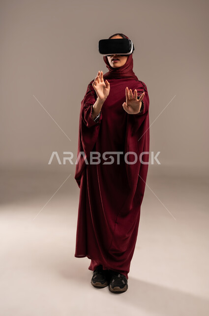 Simulating a virtual world, wearing virtual reality glasses, using a modern and advanced technical device for entertainment and amusement, a portrait of a Saudi Arabian Gulf woman wearing a hijab and a red abaya, putting 3D VR glasses on her eyes, full-length body, beige background