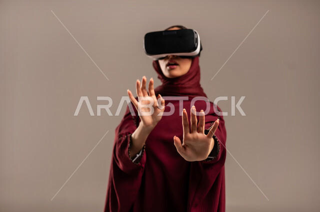 ارتداء نظارة الواقع الافتراضي، محاكاة العالم الخيالي, استخدام جهاز تقني حديث ومتطور للتسلية والترفيه, بورتريه مقرب لسيده عربية خليجية سعودية ترتدي الحجاب والعباءة الحمراء تضع نظارات الفي ار ثلاثية الأبعاد على عينها، خلفية بيج