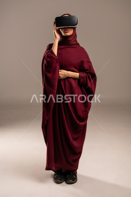 محاكاة العالم الخيالي, ارتداء نظارة الواقع الافتراضي, استخدام جهاز تقني حديث ومتطور للتسلية والترفيه, بورتريه لسيده عربية خليجية سعودية ترتدي الحجاب والعباءة الحمراء تضع نظارات الفي ار ثلاثية الأبعاد على عينها, الجسم بالطول الكامل، خلفية بيج
