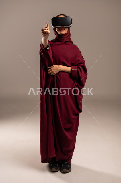 محاكاة العالم الخيالي, ارتداء نظارة الواقع الافتراضي, استخدام جهاز تقني حديث ومتطور للتسلية والترفيه, بورتريه لسيده عربية خليجية سعودية ترتدي الحجاب والعباءة الحمراء تضع نظارات الفي ار ثلاثية الأبعاد على عينها, الجسم بالطول الكامل، خلفية بيج