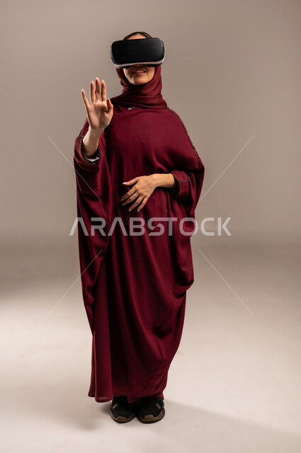 محاكاة العالم الخيالي, ارتداء نظارة الواقع الافتراضي, استخدام جهاز تقني حديث ومتطور للتسلية والترفيه, بورتريه لسيده عربية خليجية سعودية ترتدي الحجاب والعباءة الحمراء تضع نظارات الفي ار ثلاثية الأبعاد على عينها, الجسم بالطول الكامل، خلفية بيج