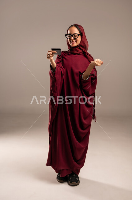 الشراء والدفع من خلال الكريدت كارد، سهولة التحويل والحساب، بورتريه لشابه عربيه خليجيه سعوديه ترتدي العباءه الحمراء والحجاب تمسك بيدها بطاقه الأتمان البنكيه تنظر اليها بإيماءات الفرح و السرور، صورة الجسم بالطول الكامل، خلفية بيج