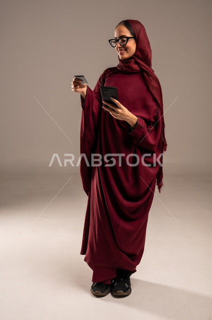 Shopping and payment through mobile programs and applications, modern and advanced technologies, portrait of a young Saudi Arabian Gulf woman wearing a red abaya and hijab holding a mobile phone in her hand, looking at a bank credit card with gestures of joy and pleasure, full-length body image, beige background