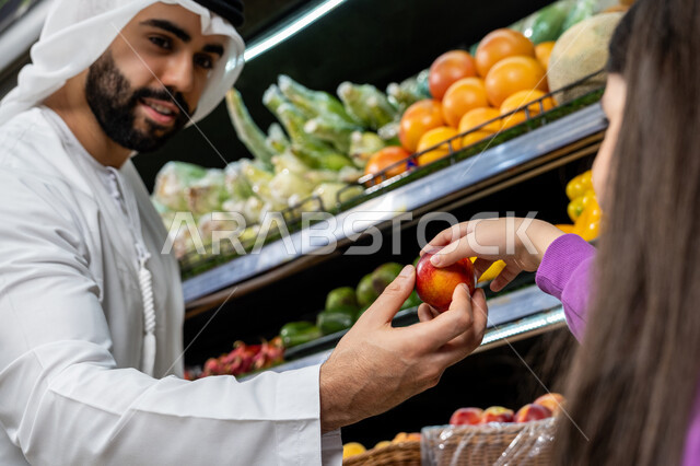 الاستمتاع بالتسوق في قسم الفواكه داخل السوبرماركت, رجل عربي خليجي اماراتي يرتدي كندورة بيضاء يقوم باعطاء ابنته ثمره تفاح طازجه, مشاركه الابناء في التسوق و شراء مستلزمات المنزل، أصناف وأنواع من مستلزمات المستهلك من مواد غذائيه، منتجات زراعيه محليه وطنيه اماراتية