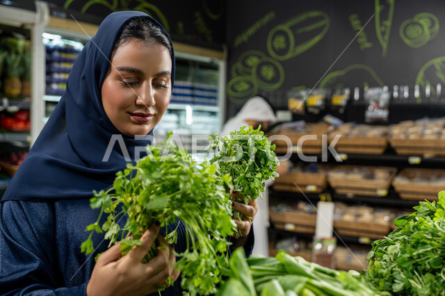 متعة التبضع واختيار الخضروات الورقية الطازجة، الاهتمام بجودة المنتجات الغذائية أثناء التسوق في السوبرماركت، امراة عربيه خليجيه اماراتية ترتدي العباءه الكحلي تقوم بالتبضع داخل قسم الخضروات، صورة مقربة لامراة تقوم باختيار البقدونس الطازج بعناية
