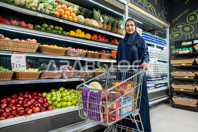 Fulfilling household needs, Saudi national agricultural crops, an Arab Gulf Emirati woman wearing an abaya shopping at the store and holding a shopping basket, a Saudi woman holding a fresh pepper in her hands and looking with gestures of joy and happiness to spend a pleasant time shopping