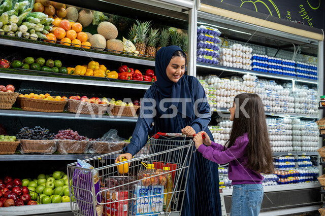 Teaching children to love helping and feel responsible from an early age, Saudi national agricultural crops, an Emirati Gulf Arab woman wearing an abaya shopping at the store, a Saudi mother picking yellow peppers and putting them in the shopping cart, gestures of joy and happiness to spend a pleasant time shopping