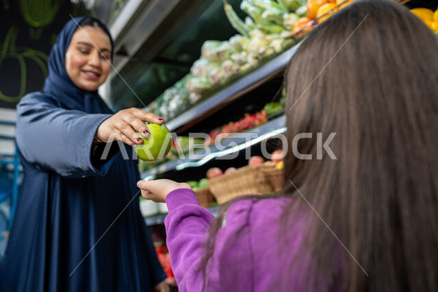 قضاء احتياجات المنزل، ايماءات الفرح والسعاده لقضاء أوقات ممتعه بالتسوق، محاصيل زراعيه وطنية سعوديه، متعة التسوق مع الأبناء، امراة عربية خليجية اماراتية ترتدي العباءة  تقوم بالتبضع من المتجر، ام سعوديه تقوم باعطاء ابنتها ثمره تفاح طازجه