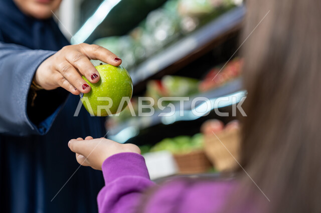 Gestures of joy and happiness for spending a pleasant time shopping, Saudi national agricultural crops, the pleasure of shopping with children, a close-up of the hand of an Arab Gulf Emirati woman wearing an abaya while shopping at the store, a Saudi mother giving her daughter a fresh apple