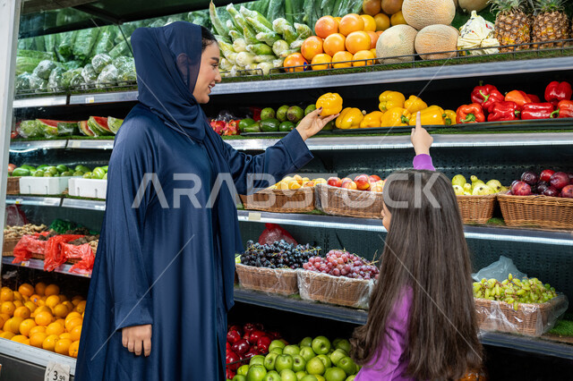 تشارك الآراء وقضاء وقت ممتع في المراكز التجارية ، متعة التسوق مع الأبناء، إمرأة عربية خليجية اماراتية مبتسمة ترتدي العباءة والحجاب تقوم بالتسوق والتبضع من السوبرماركت برفقة ابنتها، بنت سعوديه تشير إلى منتج ما في السوبرماركت