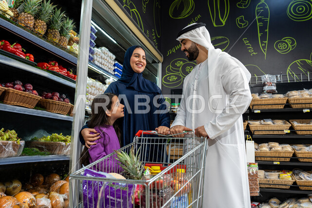 التبضع باللوازم المنزليه من قسم الخضراوات بايماءات السعاده و السرور، عائلة عربية خليجيه اماراتية تقوم بشراء المواد الغذائية داخل السوبرماركت برفقه ابنتهم، تعاون الزوجين في شراء المؤنة الغذائية، عربة تسوق تحتوي على المنتجات الغذائية، محاصيل زراعيه وطنية اماراتية