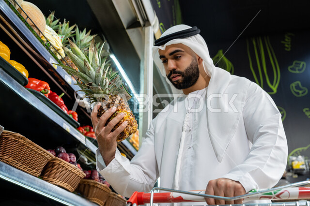 قضاء وقت ممتع في المركز التجاري, صورة مقربة لرجل عربي خليجي اماراتي  يرتدي الكندورة البيضاء يقوم بفحص ثمرة ألانانس أثناء تسوقه داخل أحد السوبرماركت , شراء الاحتياجات والمستلزمات المنزلية للمستهلك, السلع والمواد الغذائية المختلفة