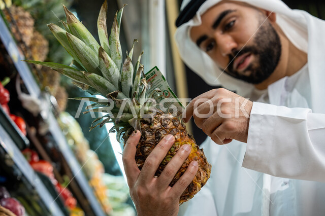 قضاء وقت ممتع في اختيار اللوازم المنزليه، مأكولات مفيدة صحية على رفوف المتجر, السلع والمواد الغذائية المختلفة, صورة مقربة لرجل عربي خليجي إماراتي يرتدي الكندورة البيضاء يقوم بفحص ثمرة ألانانس أثناء تسوقه داخل أحد السوبرماركت، محاصيل زراعيه وطنية اماراتية